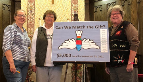 Three women standing in a church holding a sign for a $5,000 matching gift fundraising campaign.